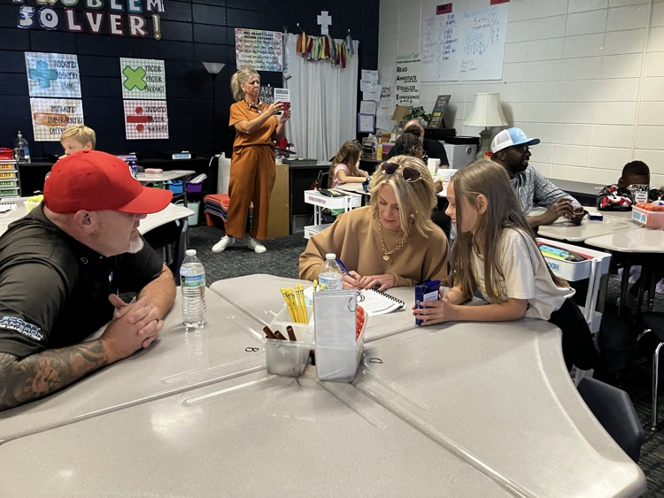 People seated at tables in a classroom, one man with a red hat, and a woman holding a microphone.