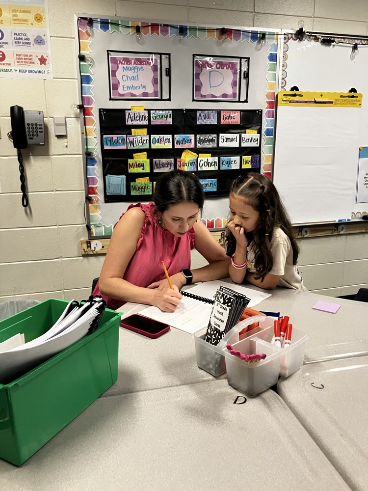Two students sitting at a desk, one writing, other holding chin with hand. Papers, pens, and other supplies on table. Whiteboard in background.