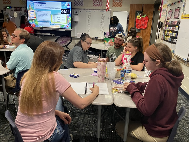 People in a classroom at tables, one writing, others looking at a monitor. A teacher stands near a whiteboard.