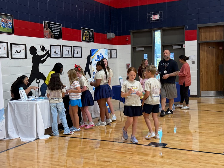 Basketball court with wooden floor. Four people at tables. Five students in cheerleading outfits stand in line.