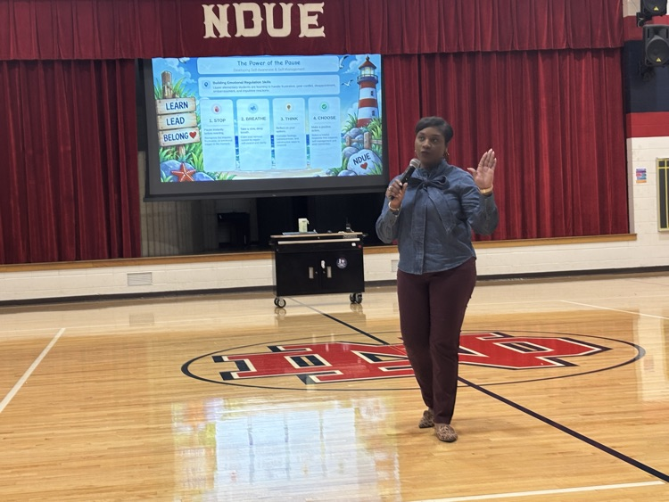 Woman stands in a gymnasium, speaking into a microphone, with a projector screen displaying a list behind her.