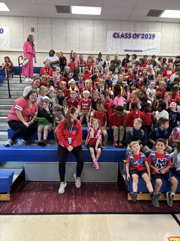A gymnasium with many students and adults seated on bleachers, some wearing red shirts.
