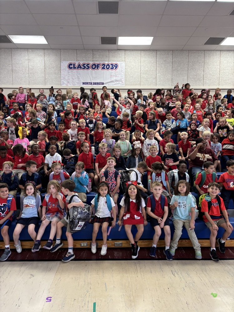 Many children seated in a large auditorium. Some wear red, others wear various colors.