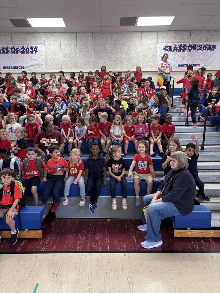 Many young students seated on bleachers in a school gym, wearing red and yellow shirts. An adult seated on the lower bleacher with students.