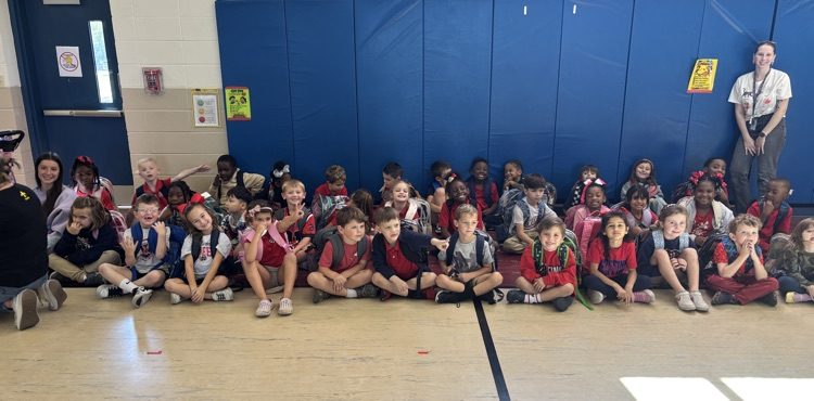 Many children in school uniforms sitting on a gym floor with an adult standing near a blue wall.