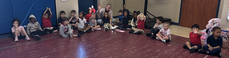 Many children seated in a circle on a red floor, some wearing hoodies, with a blue wall and door in background.