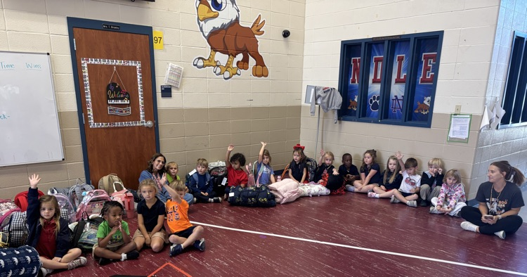 A group of students are seated on the floor in a gymnasium, some with hands raised.
