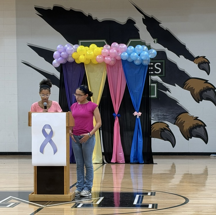 Two women standing at a podium with a ribbon and colorful balloons. One holds a microphone.