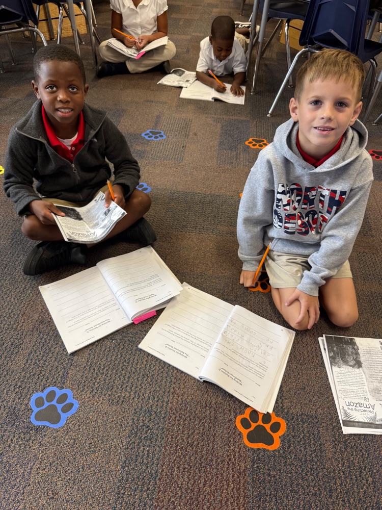 Two young boys sitting on the floor with papers and pencils. Other students sitting and reading.
