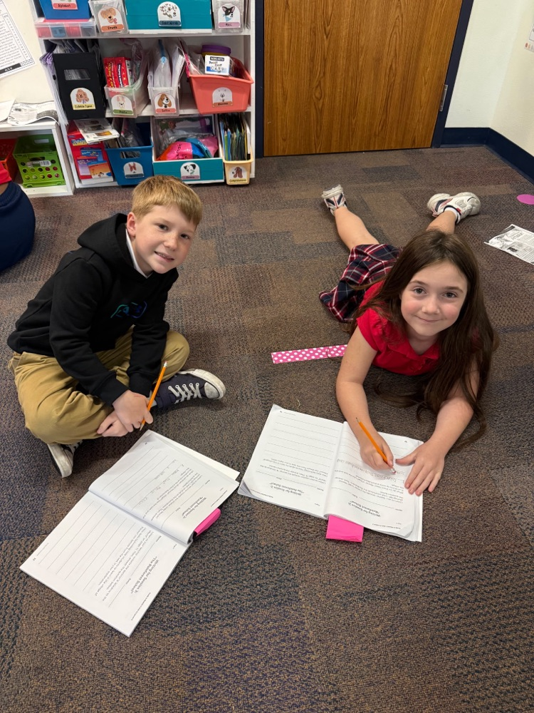 Two children work on papers while sitting on the floor; one holds a pencil, and the other uses a pen.