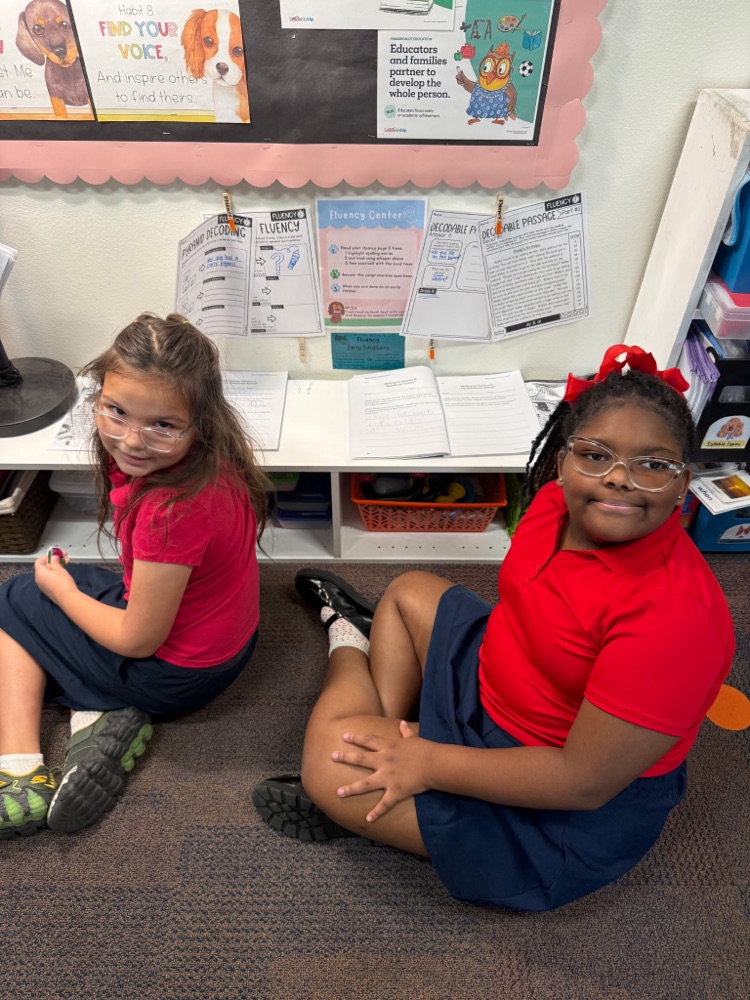 Two girls in red shirts and glasses sit on the floor in a classroom. Papers are pinned on the bulletin board.