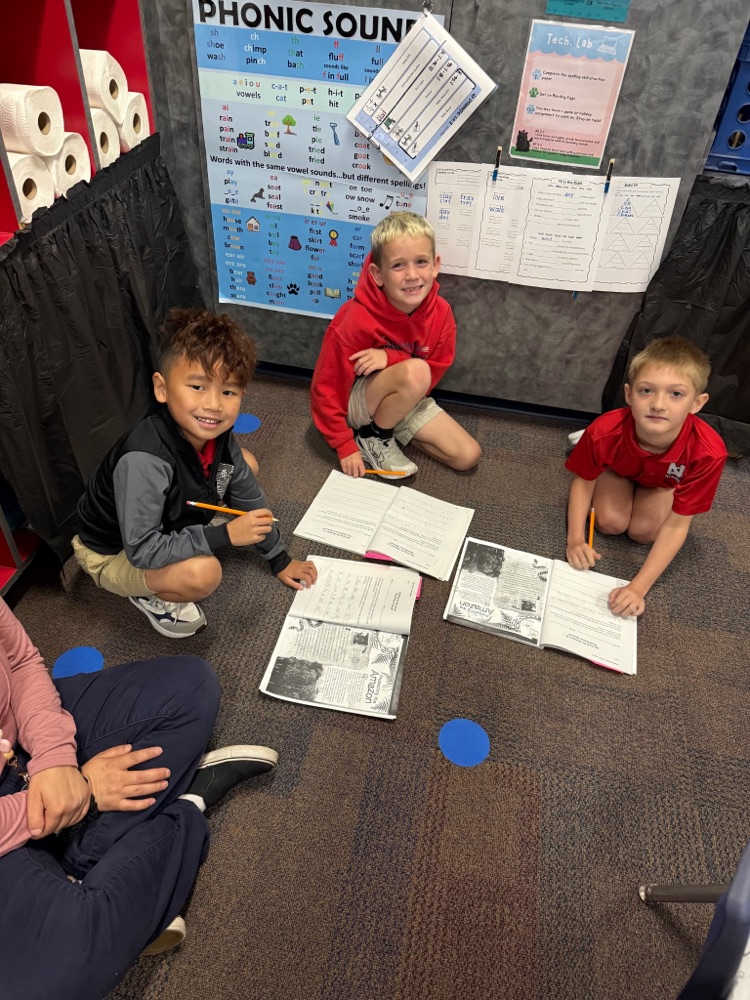 Four boys on floor with papers, pencils, and floor markers, one holding a pencil, another writing.
