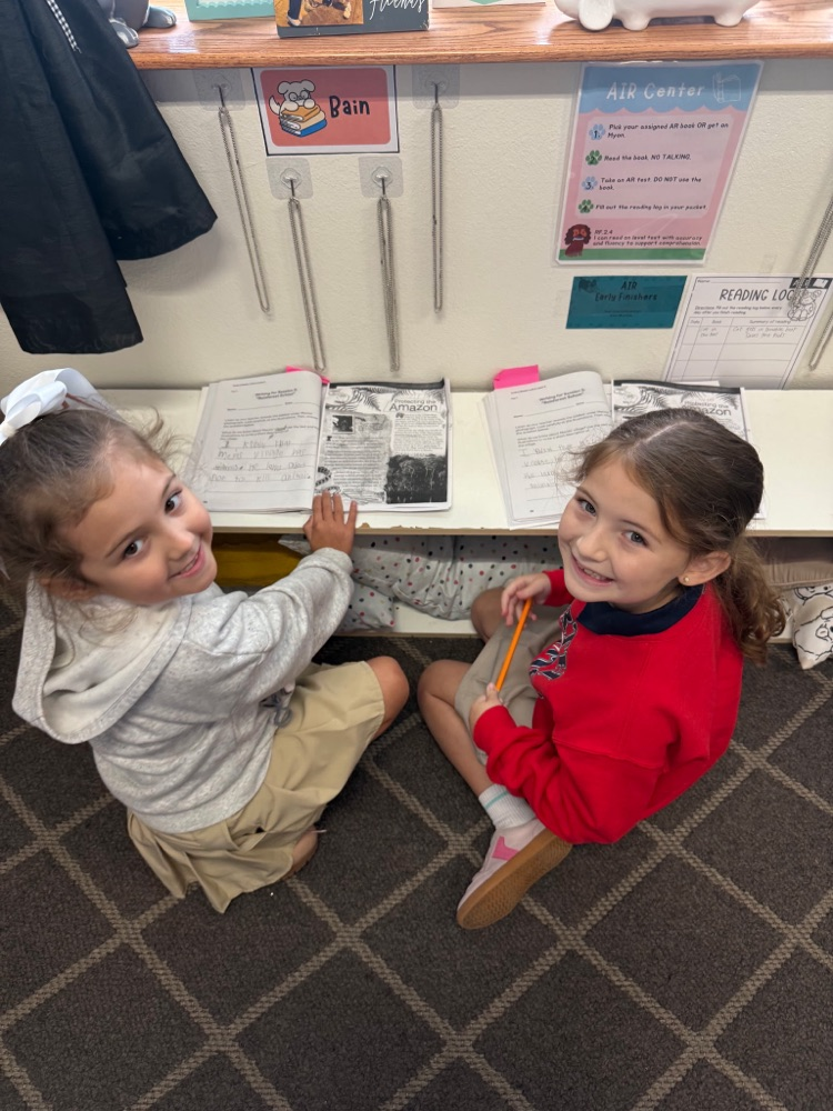 Two girls sitting on floor, one with book, the other holding pencil, smiling. Behind them, a desk with papers and a bulletin board.