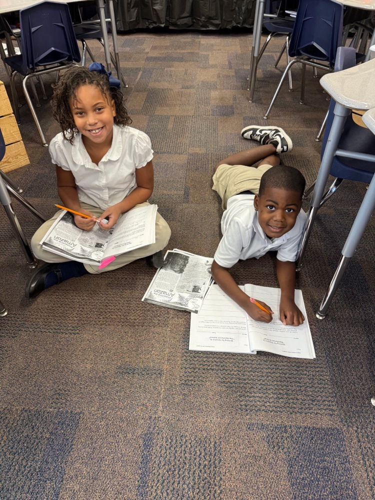 Two children sit on the floor of a classroom, both with papers and pencils. One child is seated while the other is on his stomach.