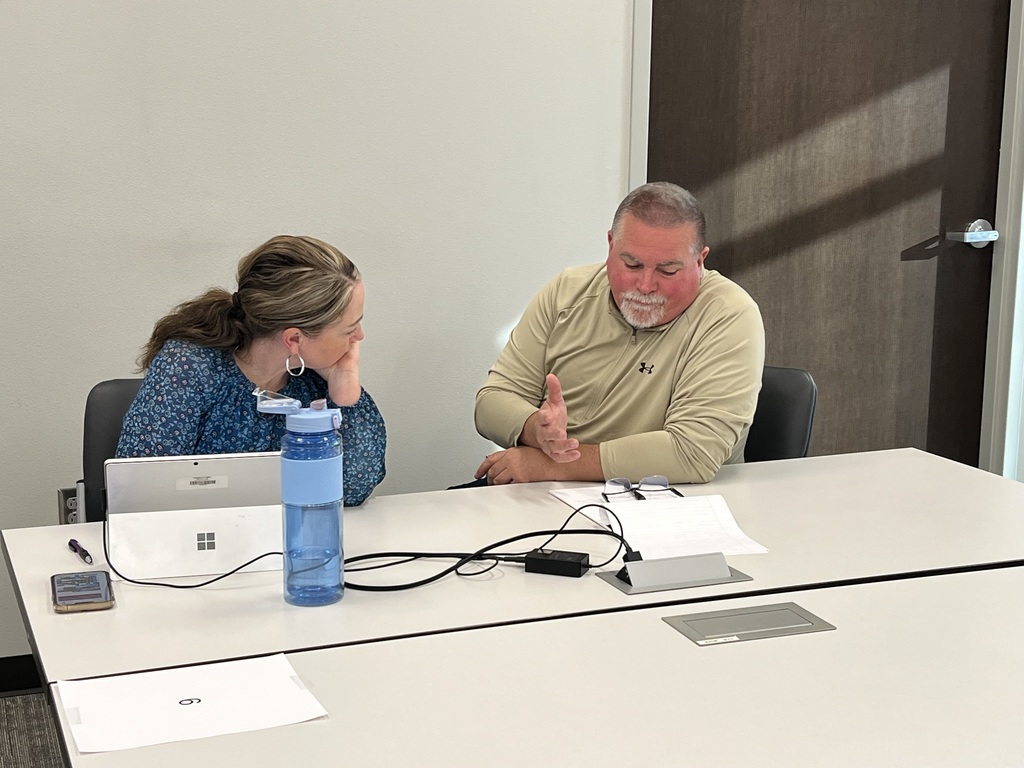 Two adults sit at a table with a laptop, water bottle, papers, and a charger.