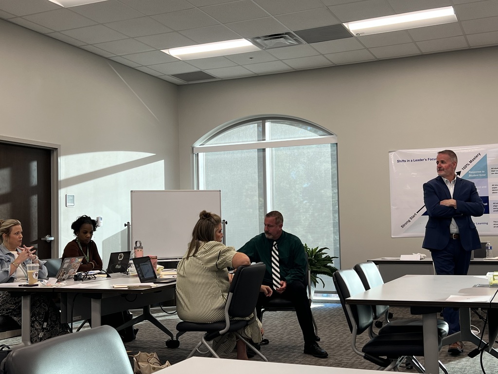 People sit in chairs around a table with laptops. A man in a suit speaks to them near a whiteboard.