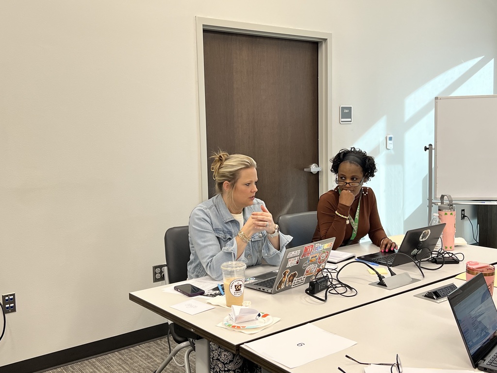 Two women seated at a table with laptops, one with a drink and the other holding a phone.