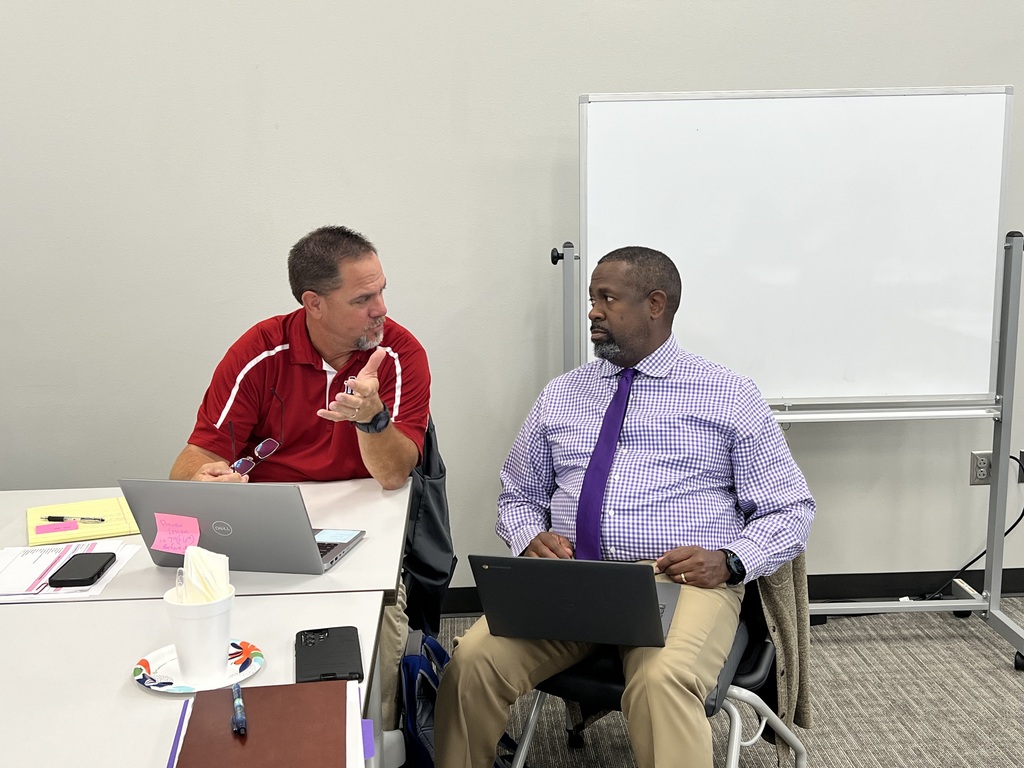 Two men in business attire sitting at a table with laptops, one gesturing, and a whiteboard in the background.