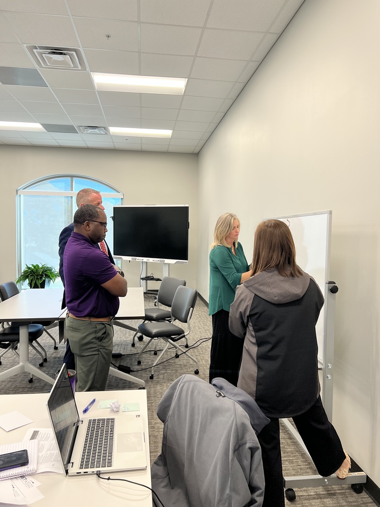 Four adults in a room, two standing by a whiteboard, another two near a table with a laptop and papers.