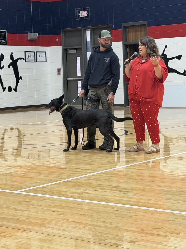A man in a cap and a woman in red pajamas stand on a basketball court with a black dog.