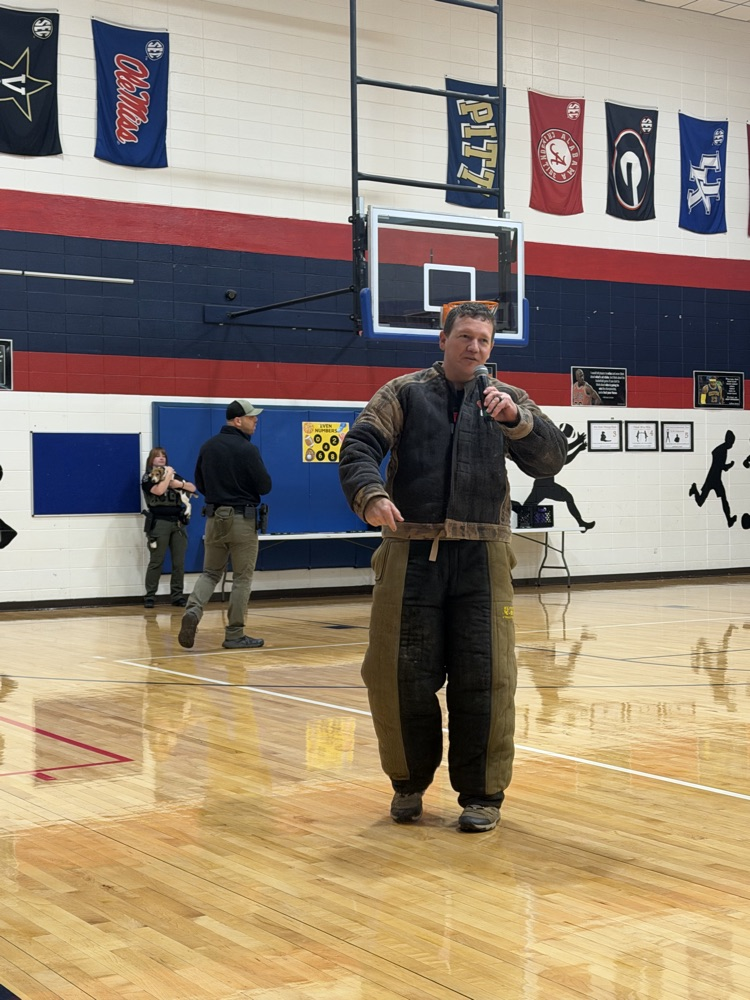 A man speaks into a microphone on a basketball court, with several people standing in the background.