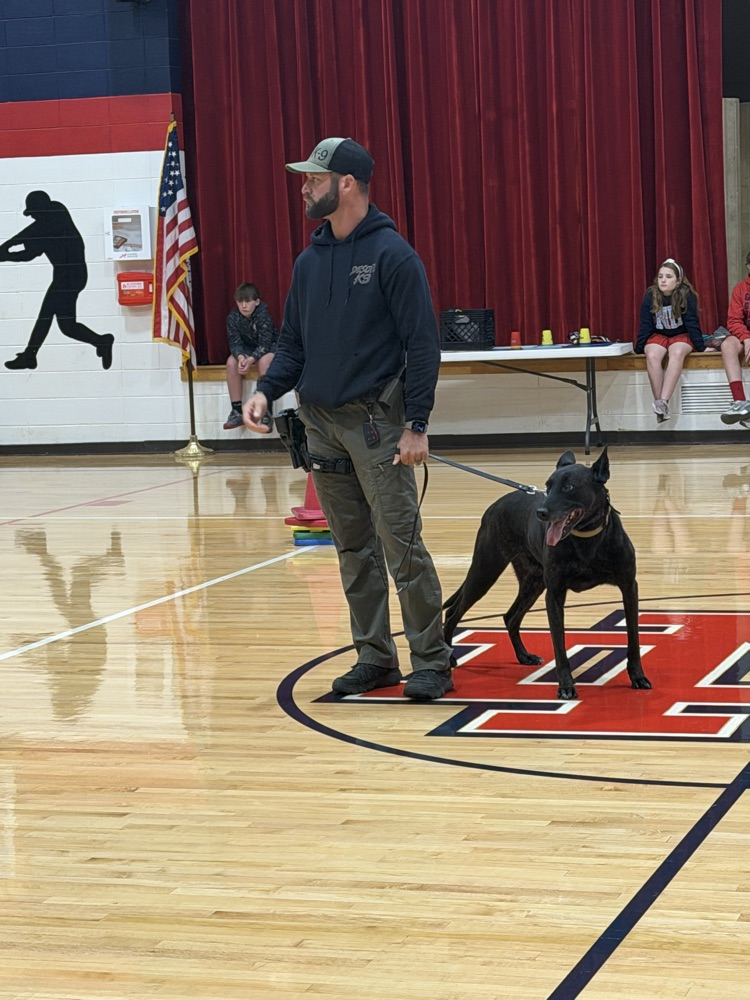 A man and dog in a gymnasium. Three people sit on a bench behind a table.
