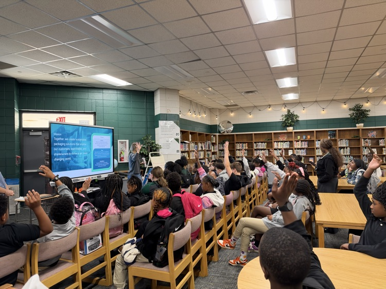 Students seated at tables in a classroom raising hands, looking at a large monitor on the wall.