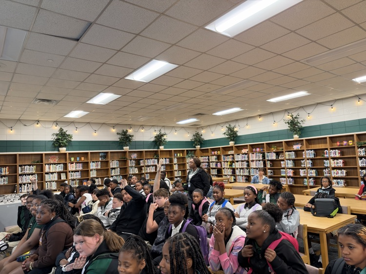 A classroom with rows of desks, some students seated and others standing, a teacher at a podium.