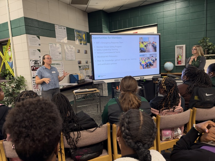 A teacher gives a presentation to students in a classroom, with a large screen displaying information.