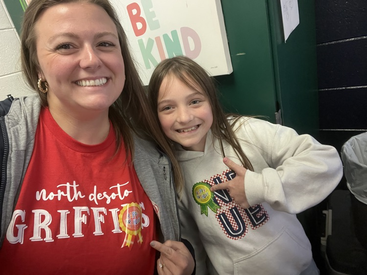 A smiling adult and child stand together; adult wears red shirt, child in white, both looking at the camera.