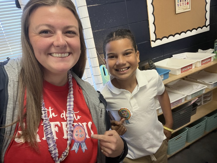 A smiling woman and child standing in a classroom, woman holding candy, child wearing a medal.