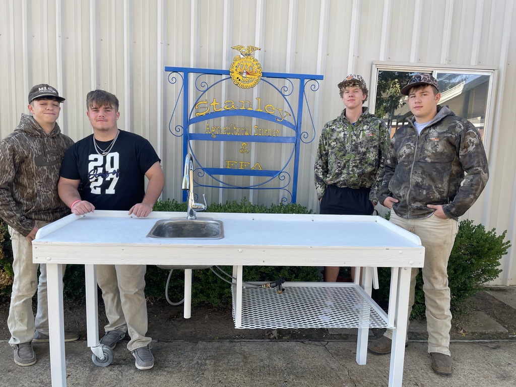 Four young men stand next to a table with a sink, posing in front of a building.
