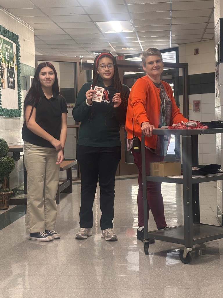 Three women stand in a hallway with a trolley. A potted plant and bench are nearby.