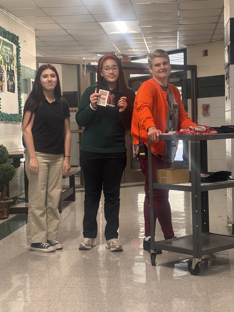 Three women stand in a hallway with a wheeled cart and one holding an object.