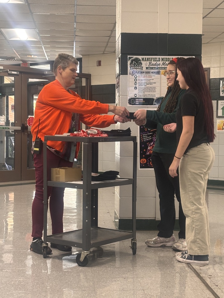 Man in orange shirt shakes hands with two women, one holding a pen, in front of a rolling cart.