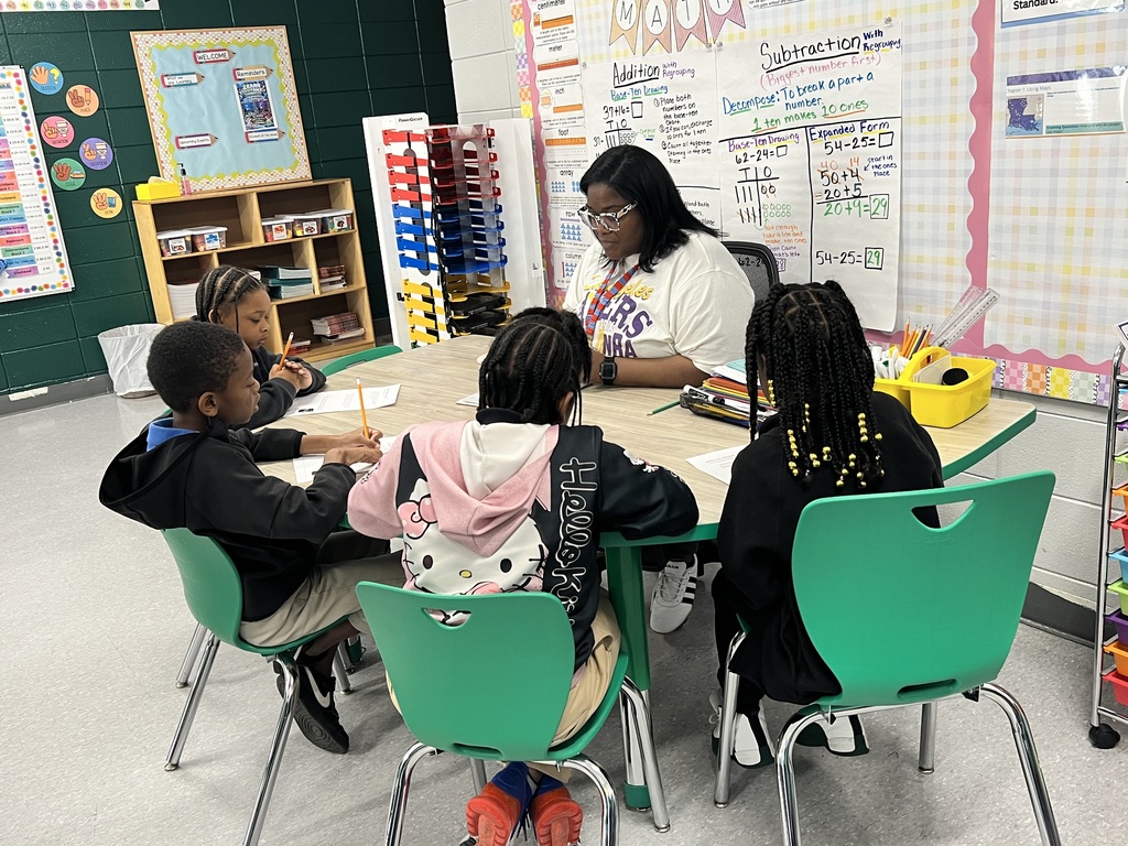Woman and four children sitting around a table with papers and pencils. Whiteboard with writing and drawings on wall.