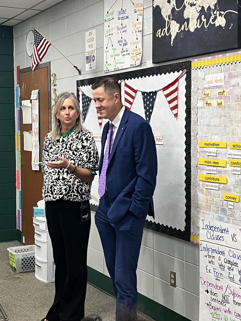 Two people stand next to bulletin board with maps, flags, and papers. Woman holds card; man looks at her.