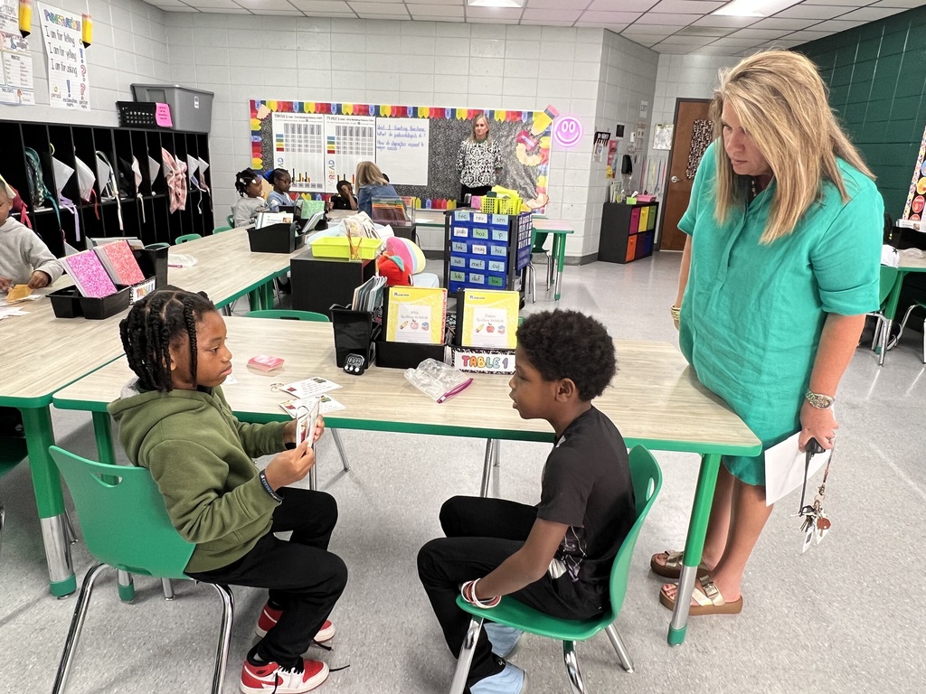 Two children seated at a desk. One girl holds a paper. The other boy looks at her. A woman in green stands behind them.