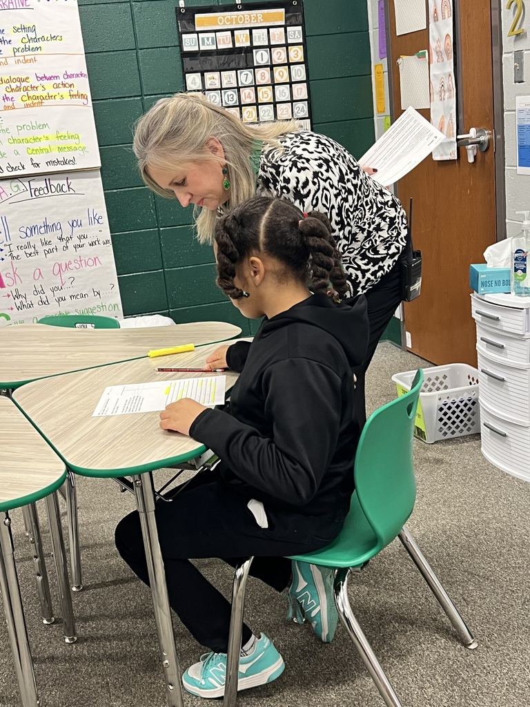 Woman leaning over desk while student writes, looking at papers. Small classroom with white walls and green chairs.