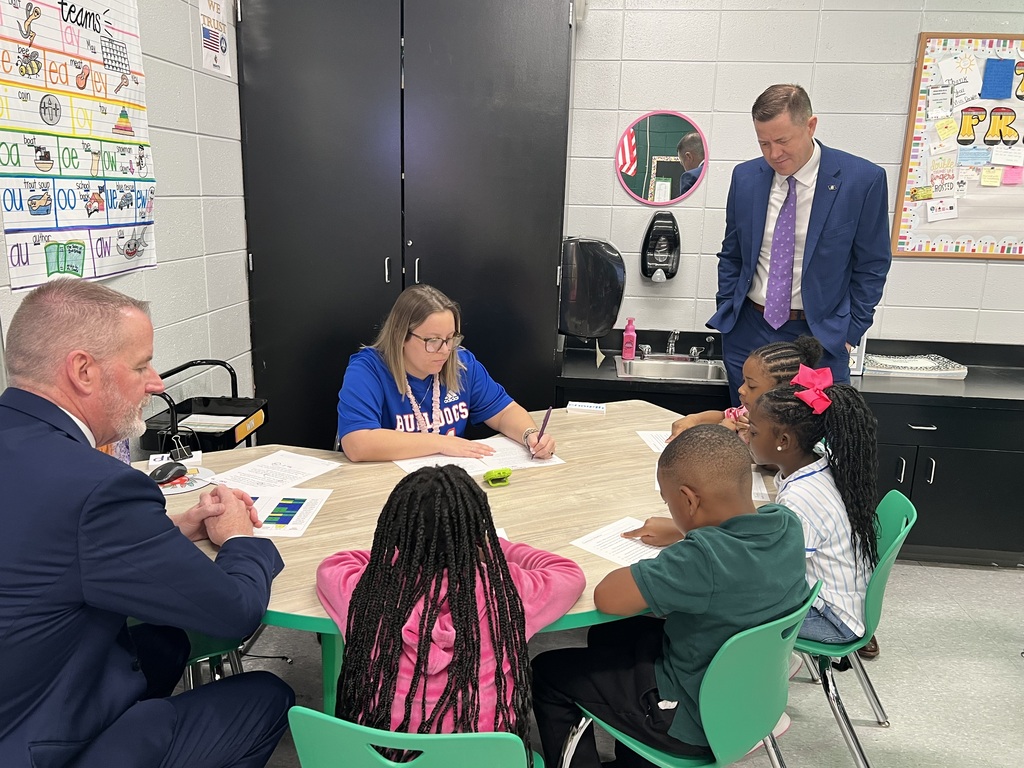 Adults and children in a classroom around a table with papers and pens, one adult writing.