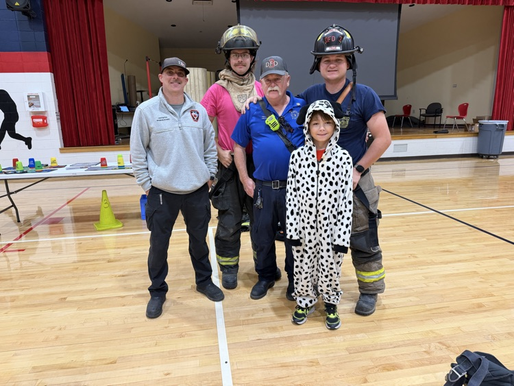 Five people stand in a gymnasium. A small child wears a spotted outfit. Three men wear blue uniforms and helmets.