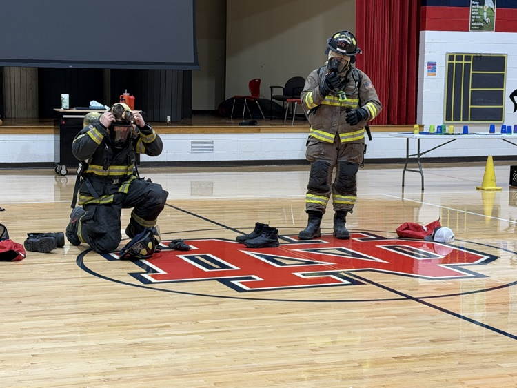 Two firefighters, one kneeling and one standing, wearing uniforms and helmets, are on a gym floor.