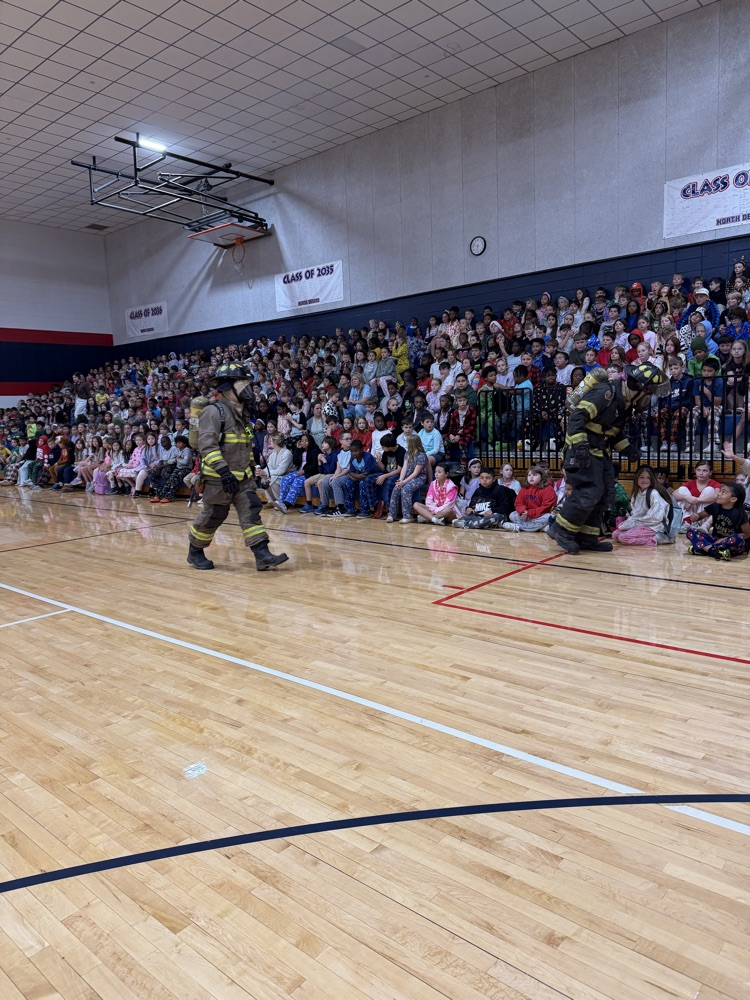Two firefighters in full uniform walk on the gym floor while many students watch from the bleachers.
