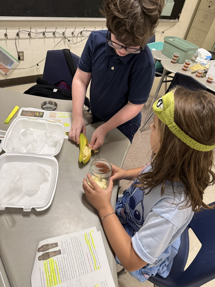 Two students work together, one holding a banana and the other holding a jar, at a table.
