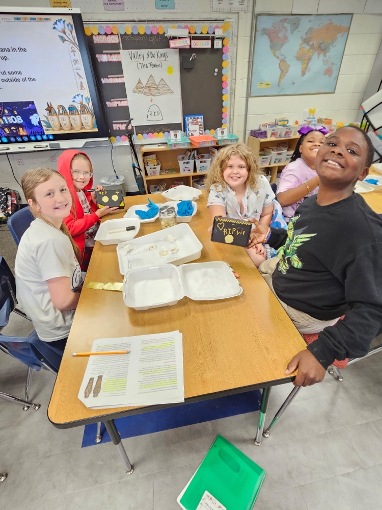 Students seated at a table with paper trays, a book, and pencils. Behind them, a classroom with a world map on the wall.