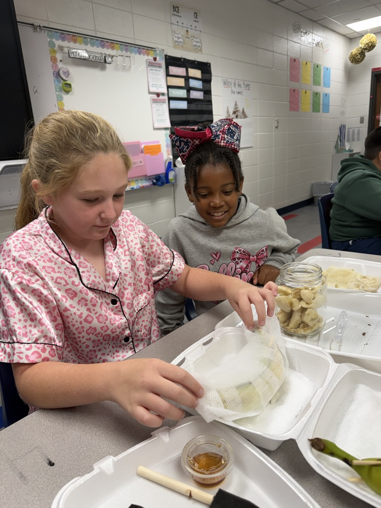 Two girls in pink and gray shirts prepare food in a classroom. One holds plastic with food, while the other holds a jar with food.