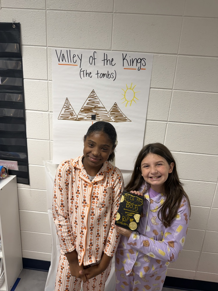 Two young girls in pajamas stand near a wall, one holding a book. Behind them, a poster says "Valley of the Kings (The Tombs)."