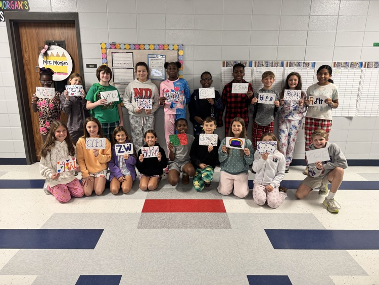Students pose with signs, some seated and others standing, in a classroom with white walls, bulletin board, and carpeted floor.
