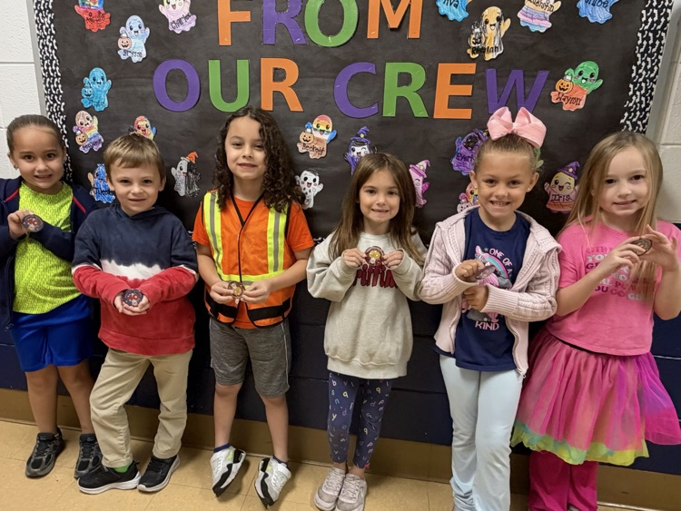 Students stand in a row in front of a black board with colorful cartoon characters and text.