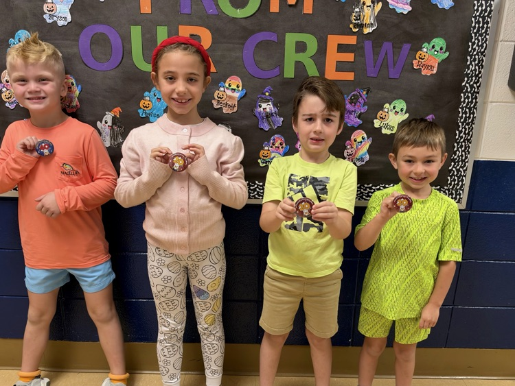 Five children stand next to each other, holding small objects, in front of a bulletin board.